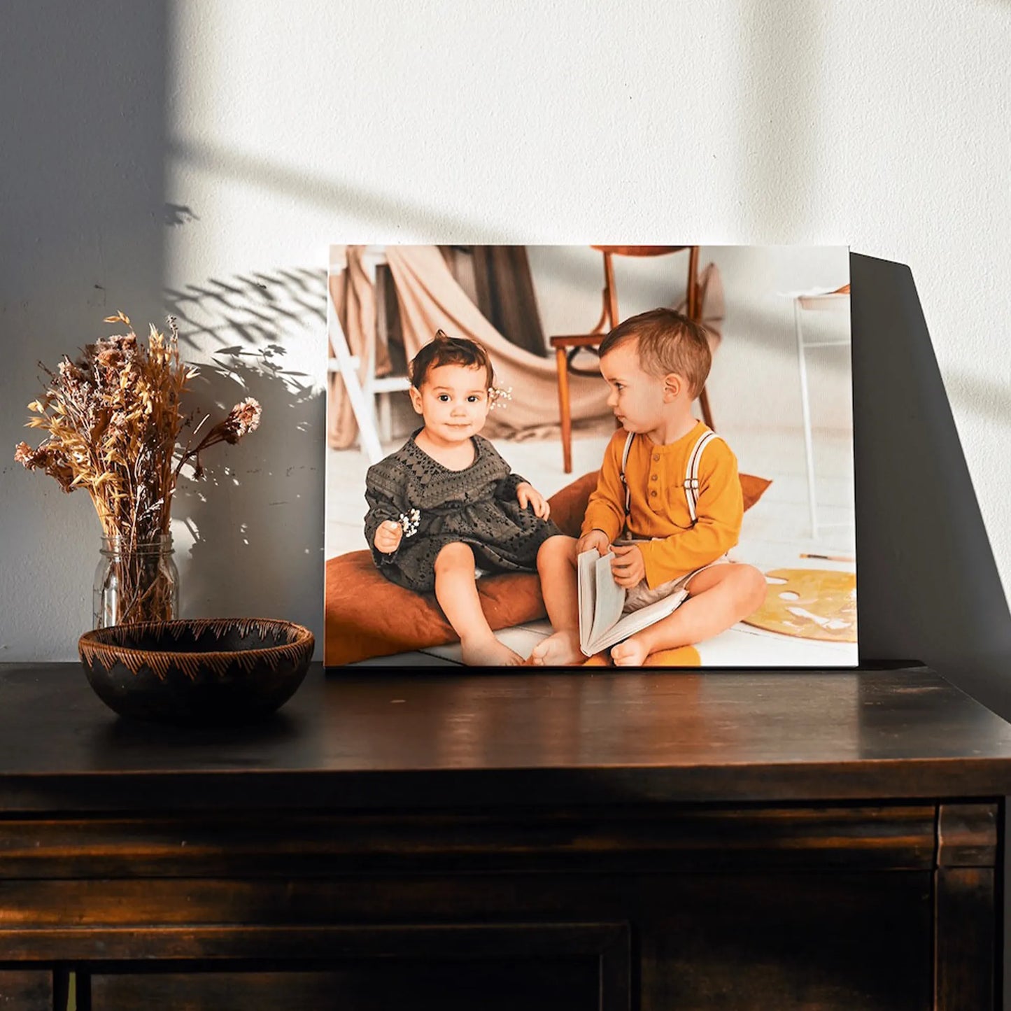 Gallery wrapped canvas print of two toddlers sitting together with a book, displayed on a dark wood console beside dried flowers.
