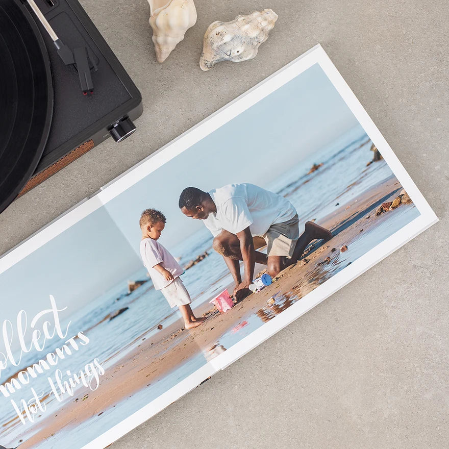 Layflat photo book open to a panoramic beach scene with a parent and child playing in the sand, placed near a record player.
