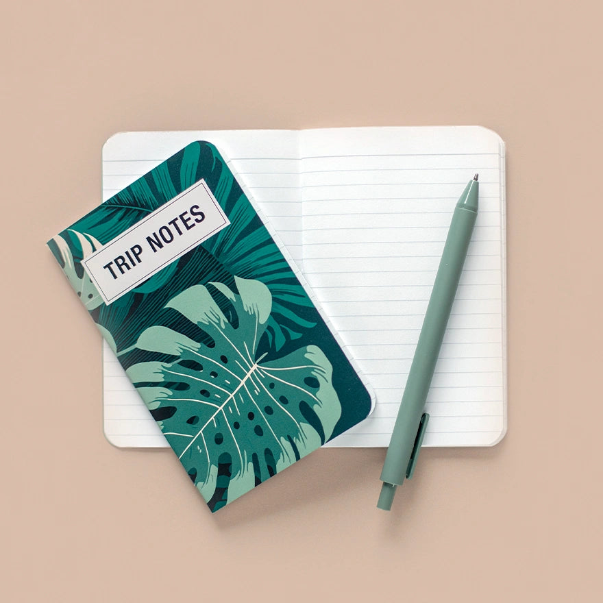 Small memo notebook with tropical leaf cover design labeled “Trip Notes,” shown open with lined pages and a pen on a neutral surface.