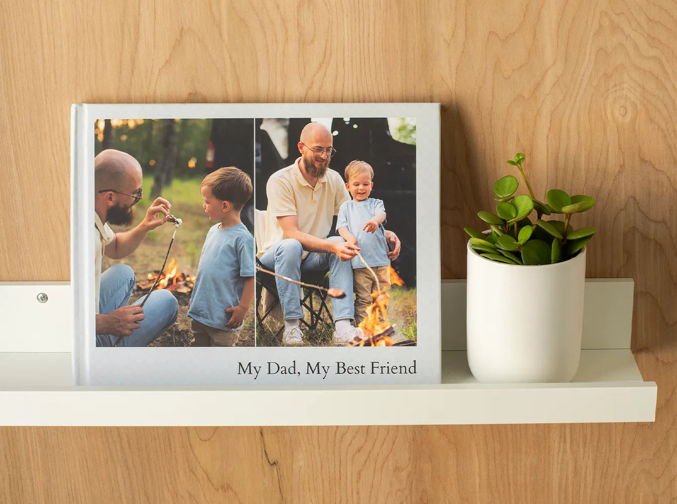 Hardcover photo book with printed cover showing a parent and child by a campfire, displayed on a shelf next to a potted plant.