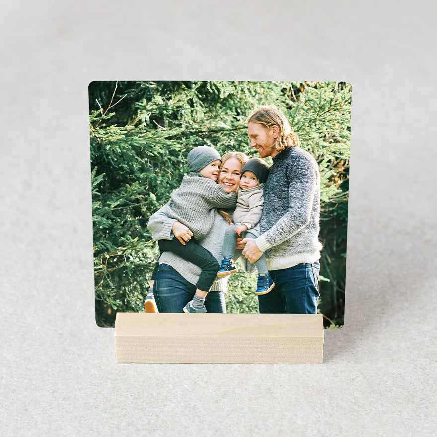 Metal desk print with rounded corners displayed in a wooden stand, featuring a family of four outdoors against evergreen trees.