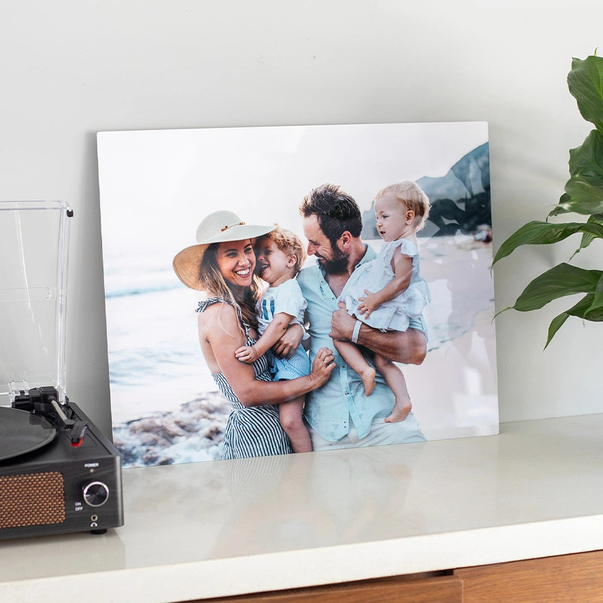 Metal wall print with glossy finish showing a family of four smiling at the beach, displayed on a shelf beside a record player and plant.