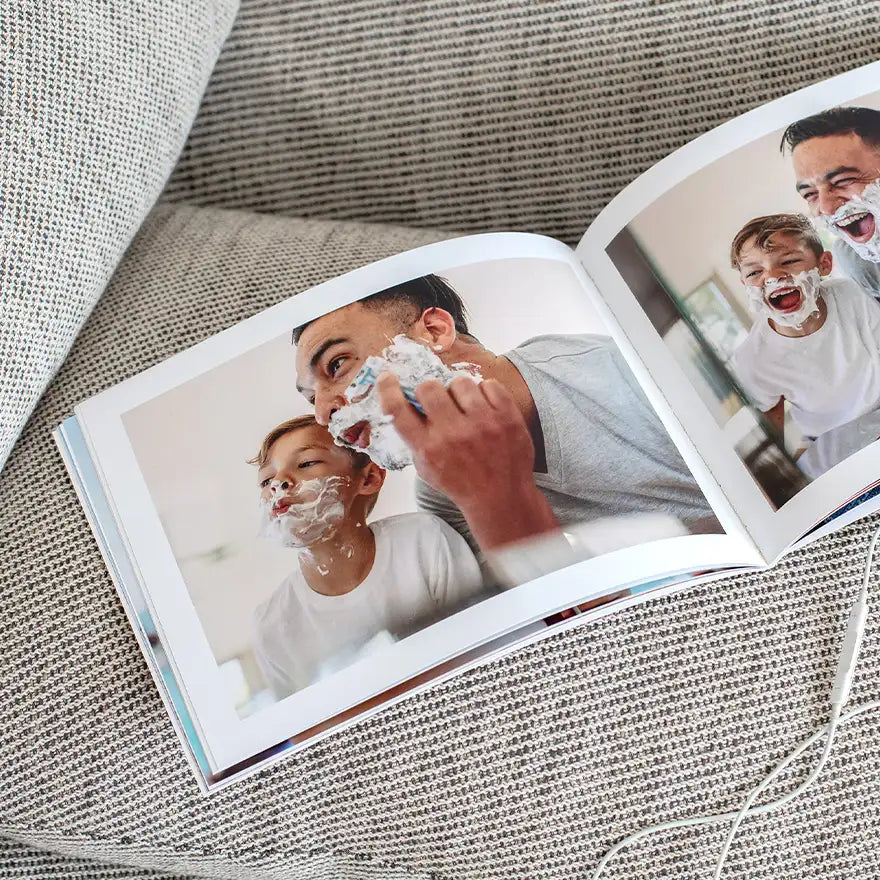 Softcover photo book open on a textured surface showing a parent and child with shaving cream on their faces in a candid layout.