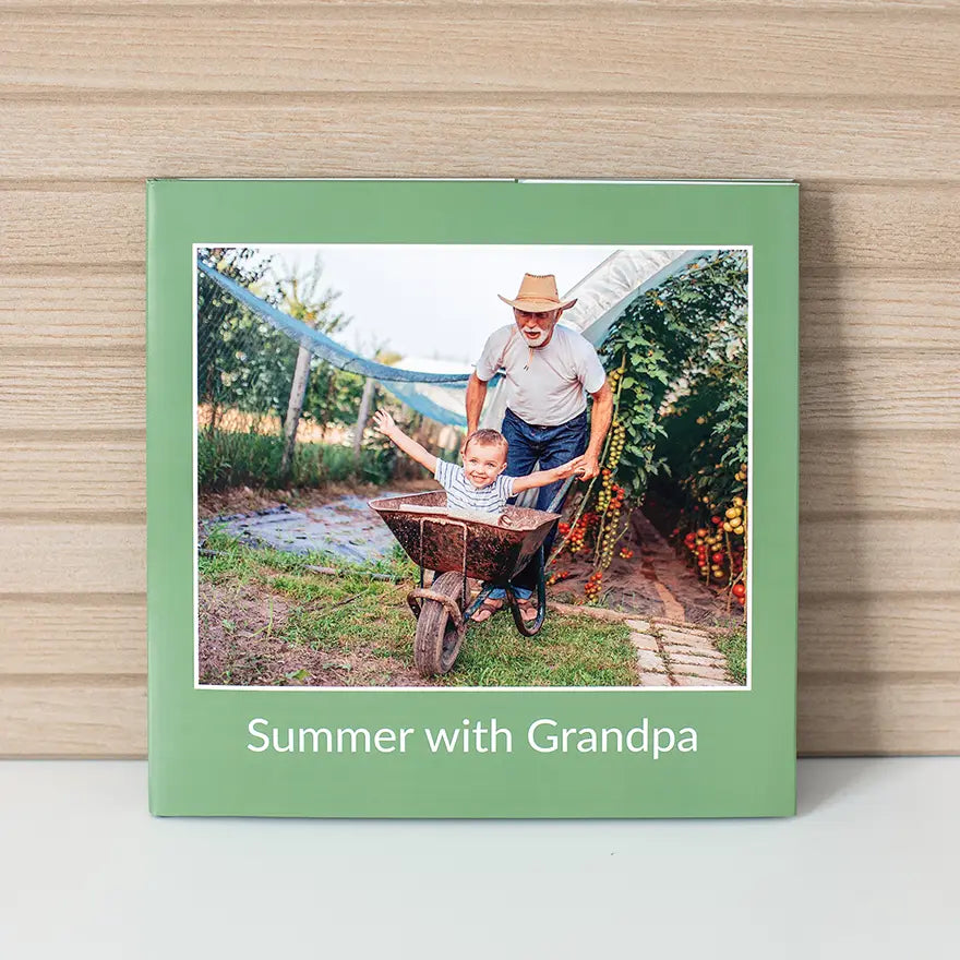 Hardcover photo book with green cover featuring a child riding in a wheelbarrow pushed by an older adult, with title text on cover.