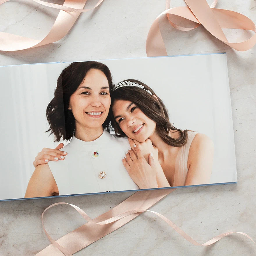 Layflat photo book open to a seamless full-bleed spread of a smiling mother and daughter with soft ribbon details around it.