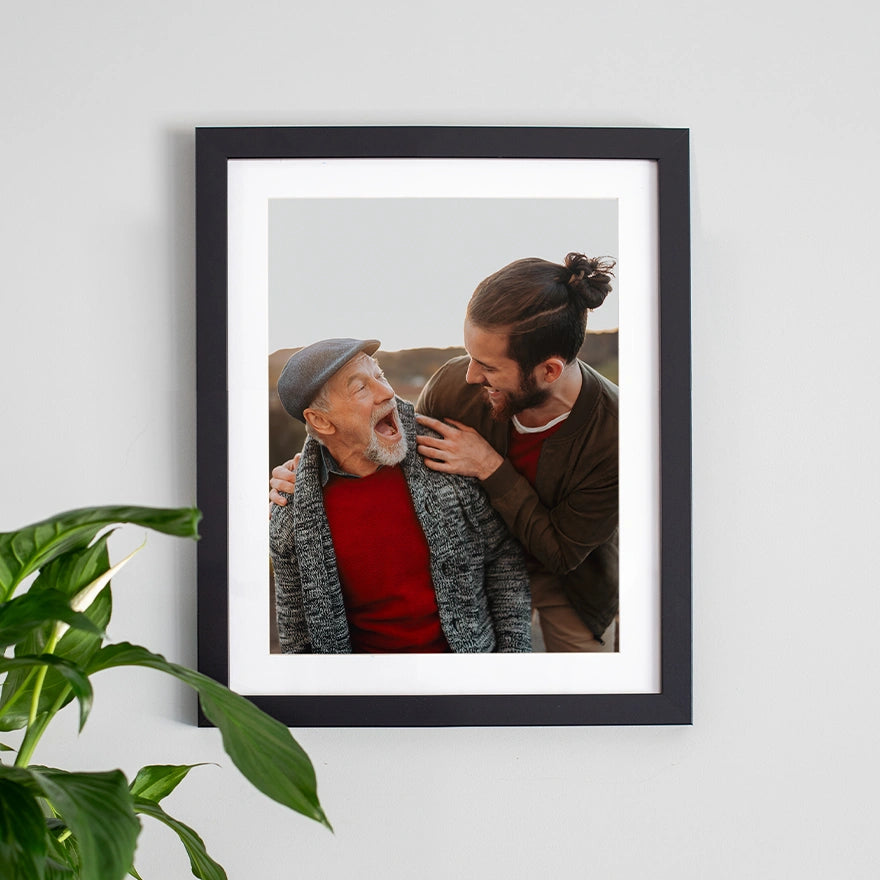 Framed photo print with white mat showing father and adult son laughing together, displayed on a wall with plant nearby
