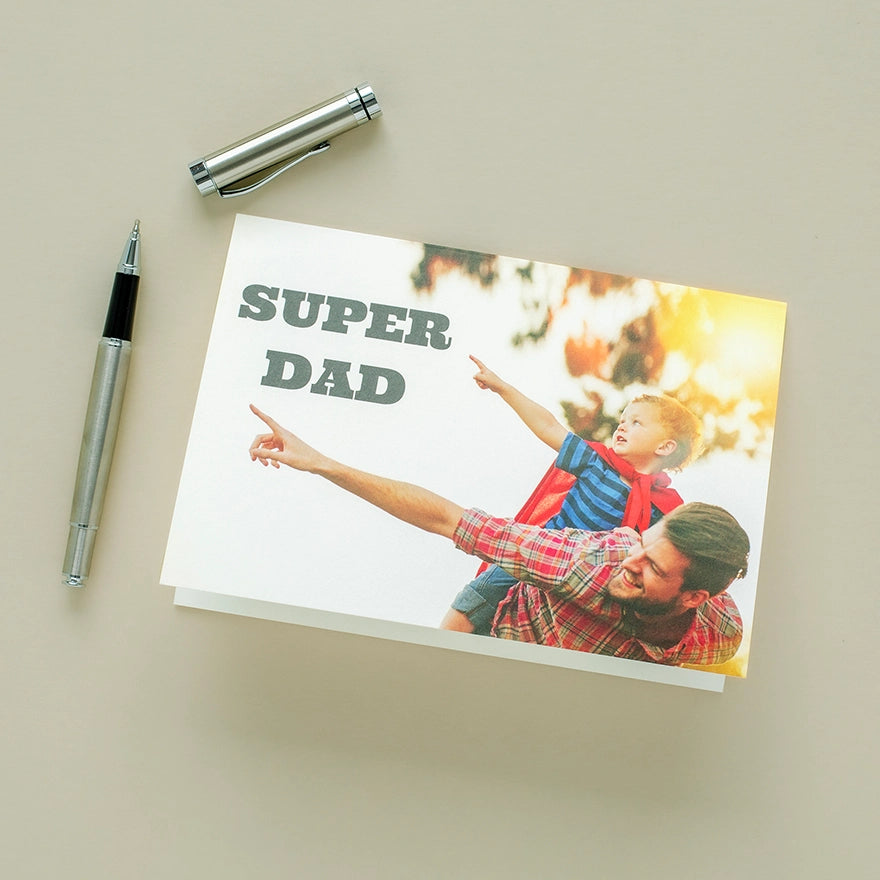 Folded photo card with “Super Dad” text and image of a father and child pointing, placed on a desk with pen