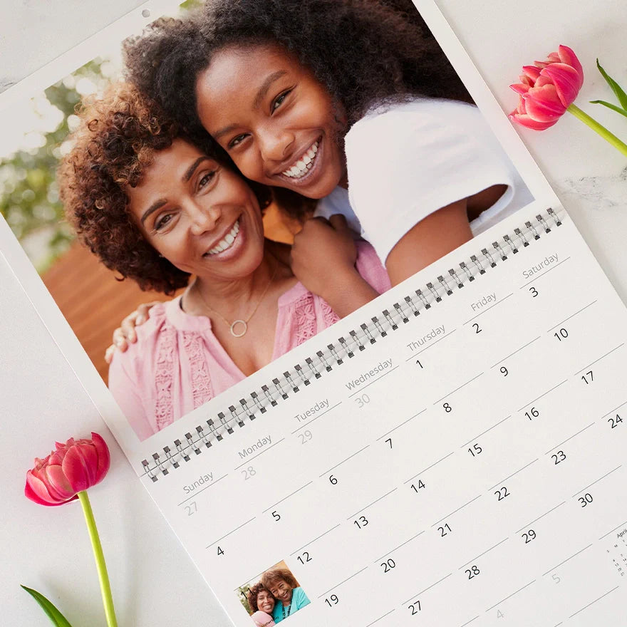 Open wall calendar with a large photo of a mother and daughter smiling together, above a clean monthly calendar layout, with pink flowers nearby.