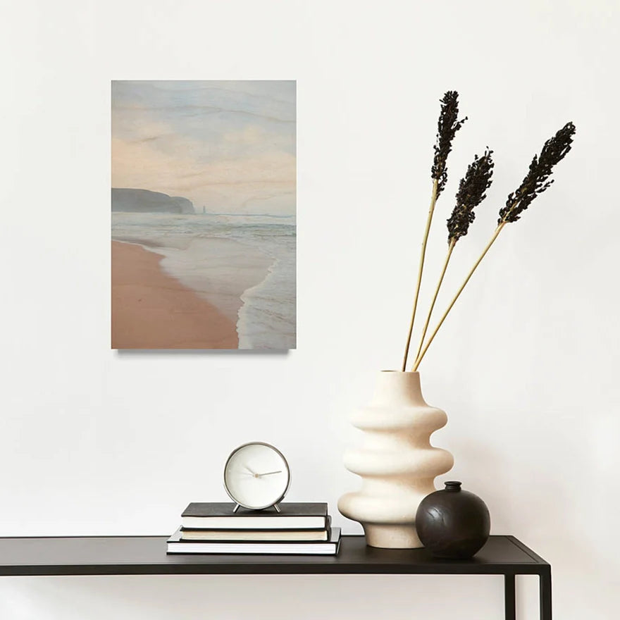 A custom wood wall print displayed above a dark console table, featuring a serene beach scene with soft pastel tones — gentle waves rolling onto sandy shores beneath a hazy, peachy sunset sky with distant cliffs. 