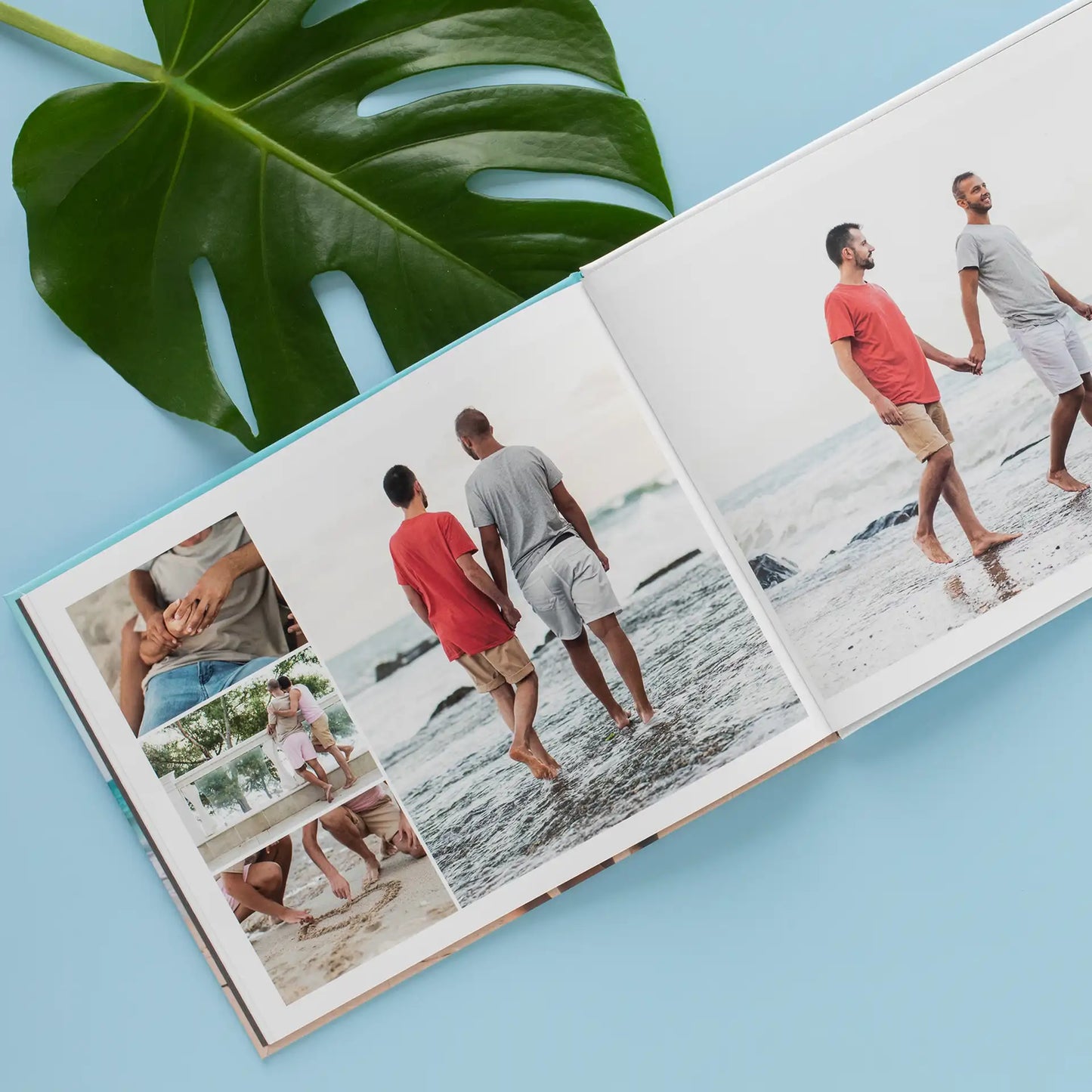 Hardcover photo book open to layout featuring couple walking on beach alongside smaller inset images, placed on blue surface with large green leaf.