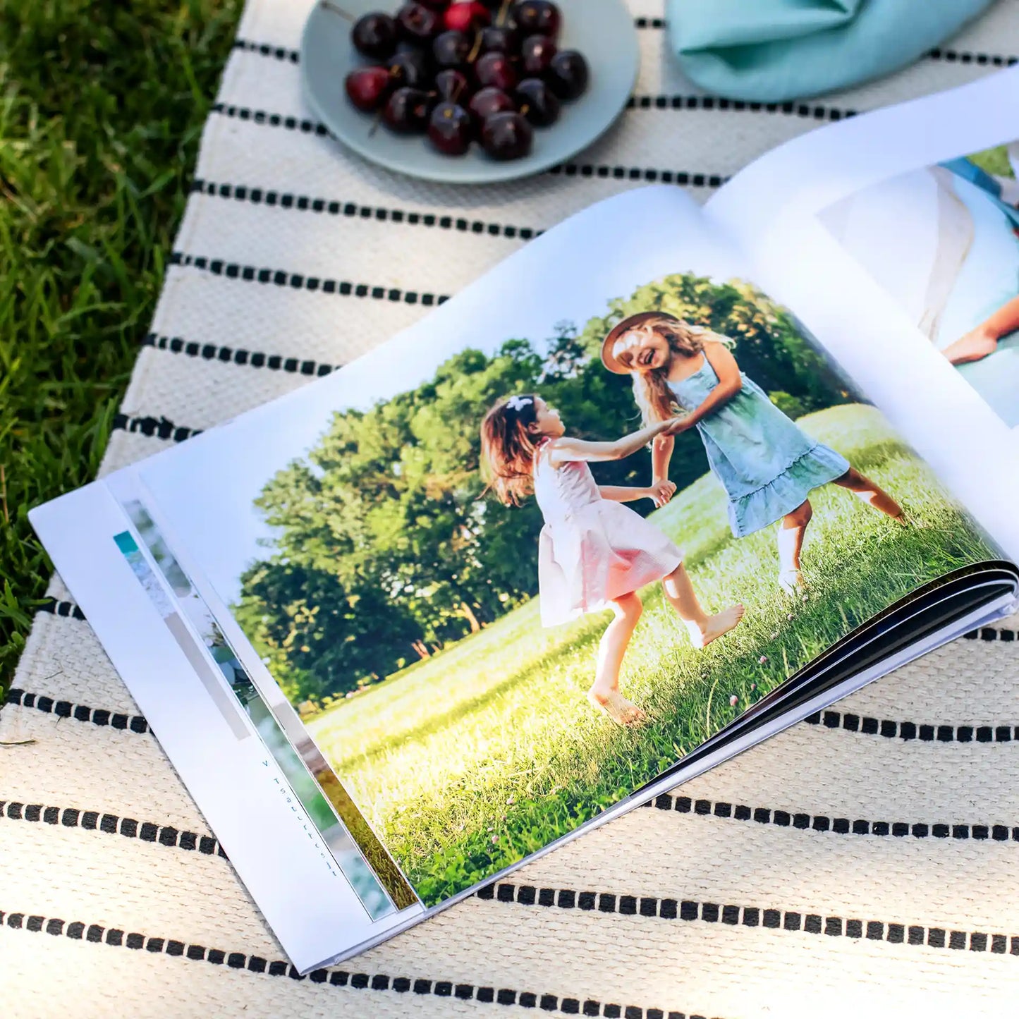 Hardcover photo book open to full-page image of two children playing on grass, displayed on picnic blanket with plate of cherries nearby.