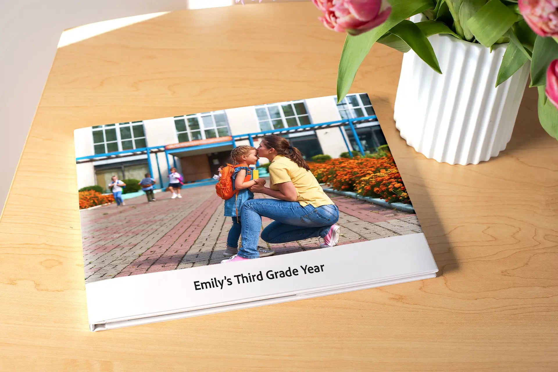 Hardcover photo book titled 'Emily's Third Grade Year' resting on a wooden table beside a white vase of pink tulips, with a cover photo of a mother sharing a tender moment with her daughter at school drop-off
