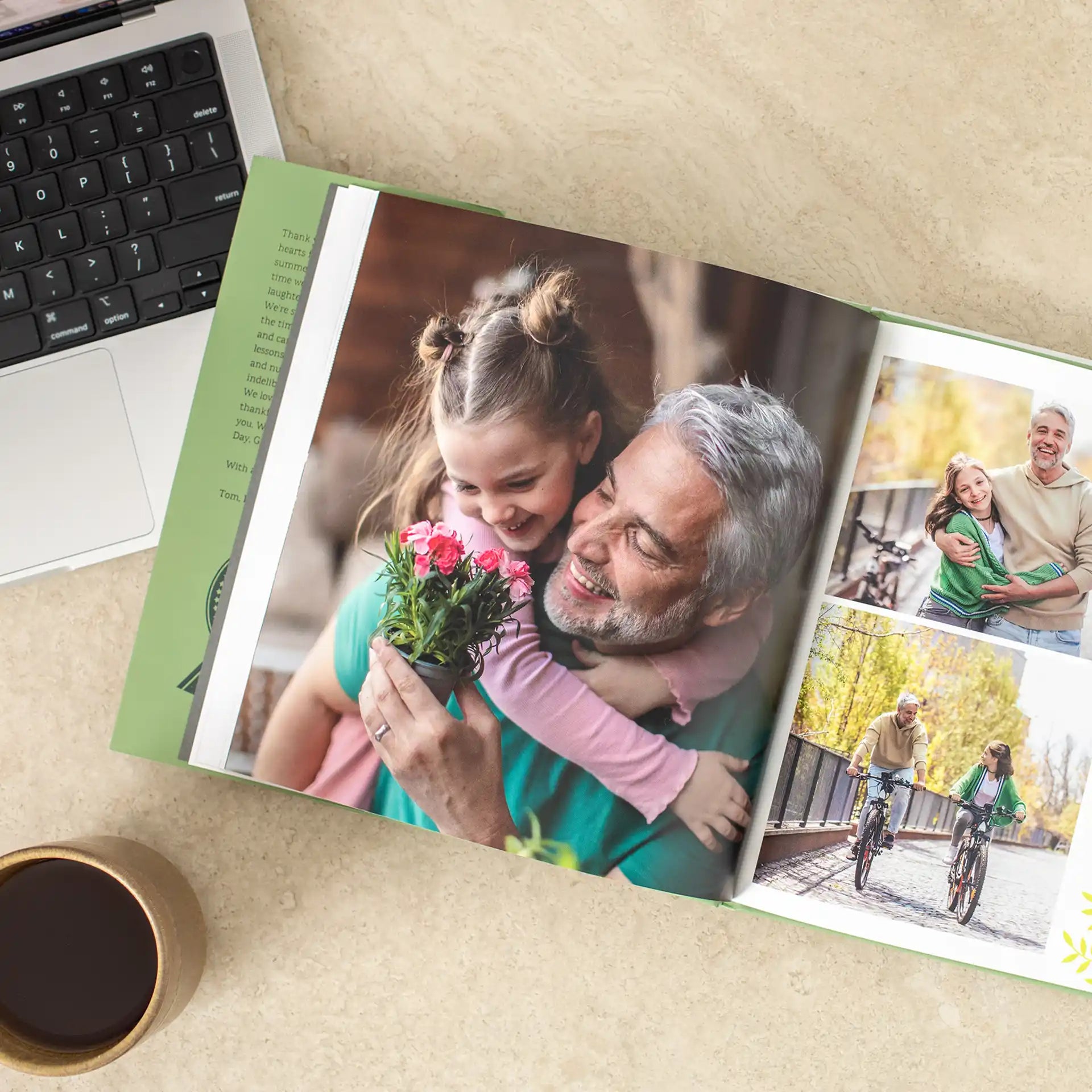 Open hardcover photo book on a desk beside a laptop and coffee cup, showing a spread featuring a large photo of a grandfather hugging his laughing granddaughter, alongside smaller photos of the pair cycling together outdoors