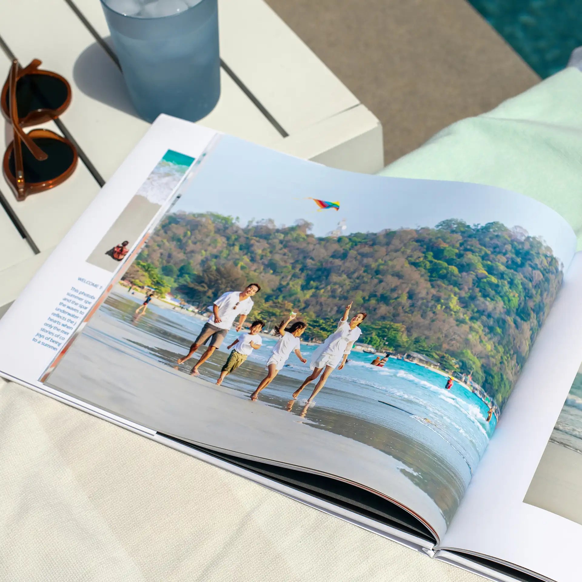 Hardcover photo book open to a full-bleed beach family photo, resting poolside beside sunglasses and a drink.