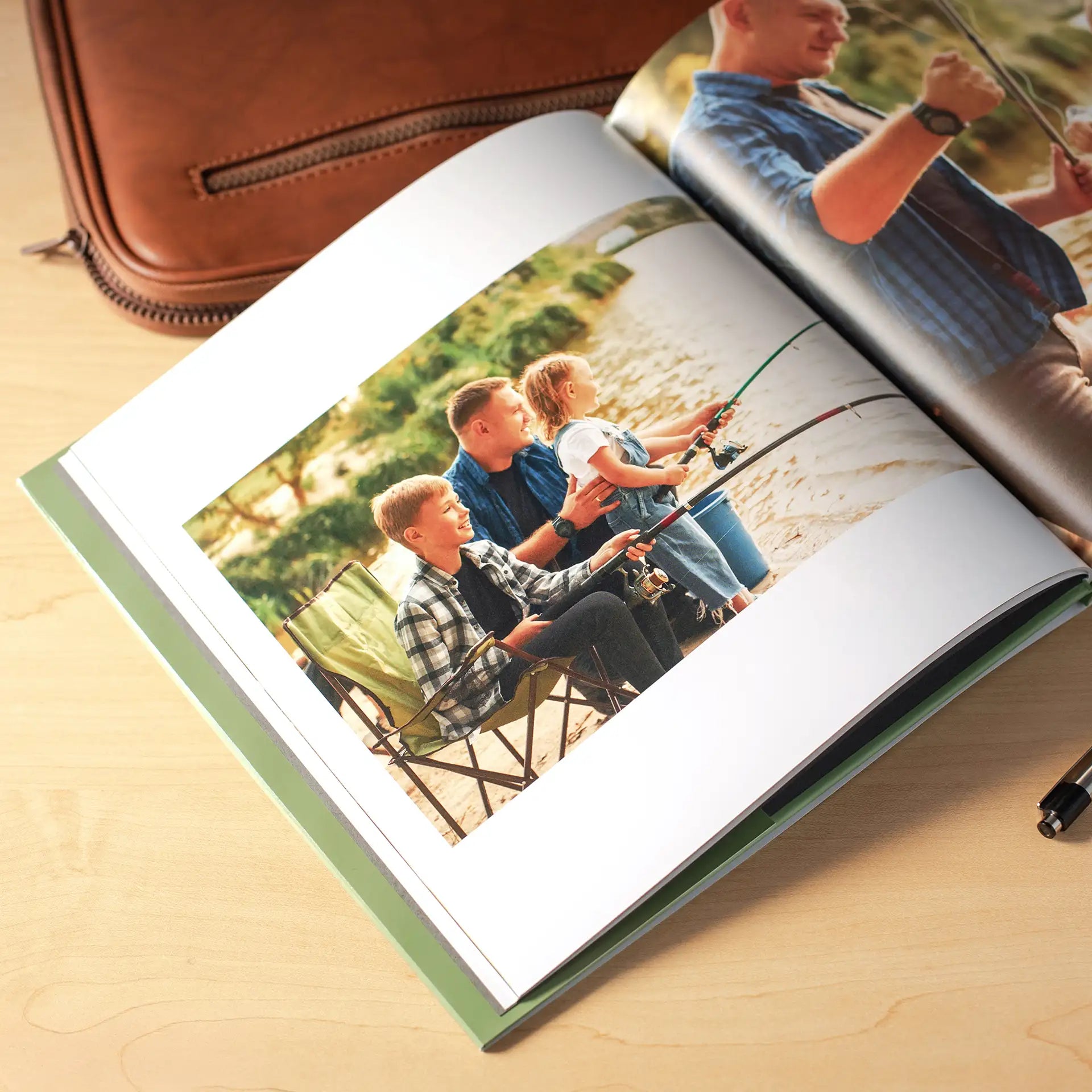 Open hardcover photo book with a green spine on a wooden surface beside a leather bag and pen, displaying a full-page spread of a father and his two young children fishing together by the water with camp chairs and fishing rods