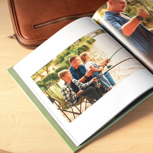 Open hardcover photo book with a green spine resting on a wooden surface beside a brown leather bag, displaying a full-page photo of a father and his two children fishing together by the water with fishing rods and camp chairs