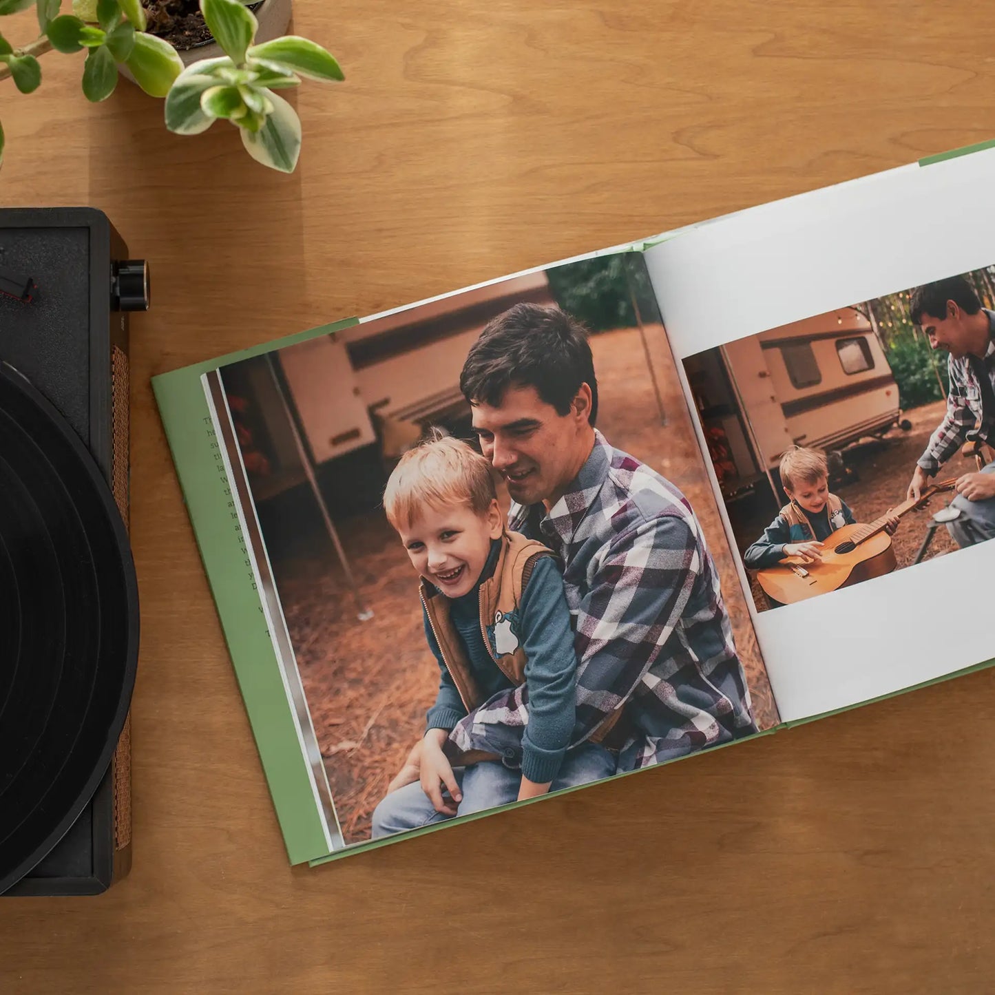 Open hardcover photo book with a green spine resting on a wooden table next to a record player and houseplant, displaying a large photo of a father and young son laughing together at a campsite, with a second photo of the boy learning to play guitar