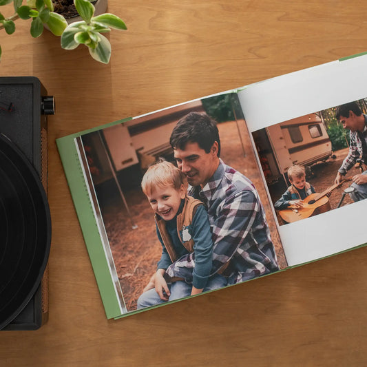 Open hardcover photo book with a green spine resting on a wooden table next to a record player and houseplant, displaying a large photo of a father and young son laughing together at a campsite, with a second photo of the boy learning to play guitar