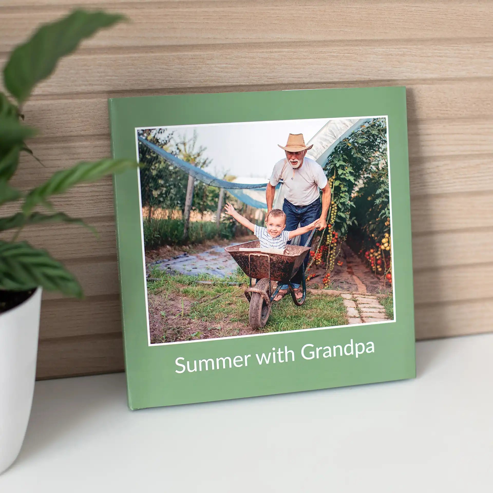 Hardcover photo book with green cover showing grandparent pushing child in wheelbarrow, with printed title text, displayed on shelf beside plant.