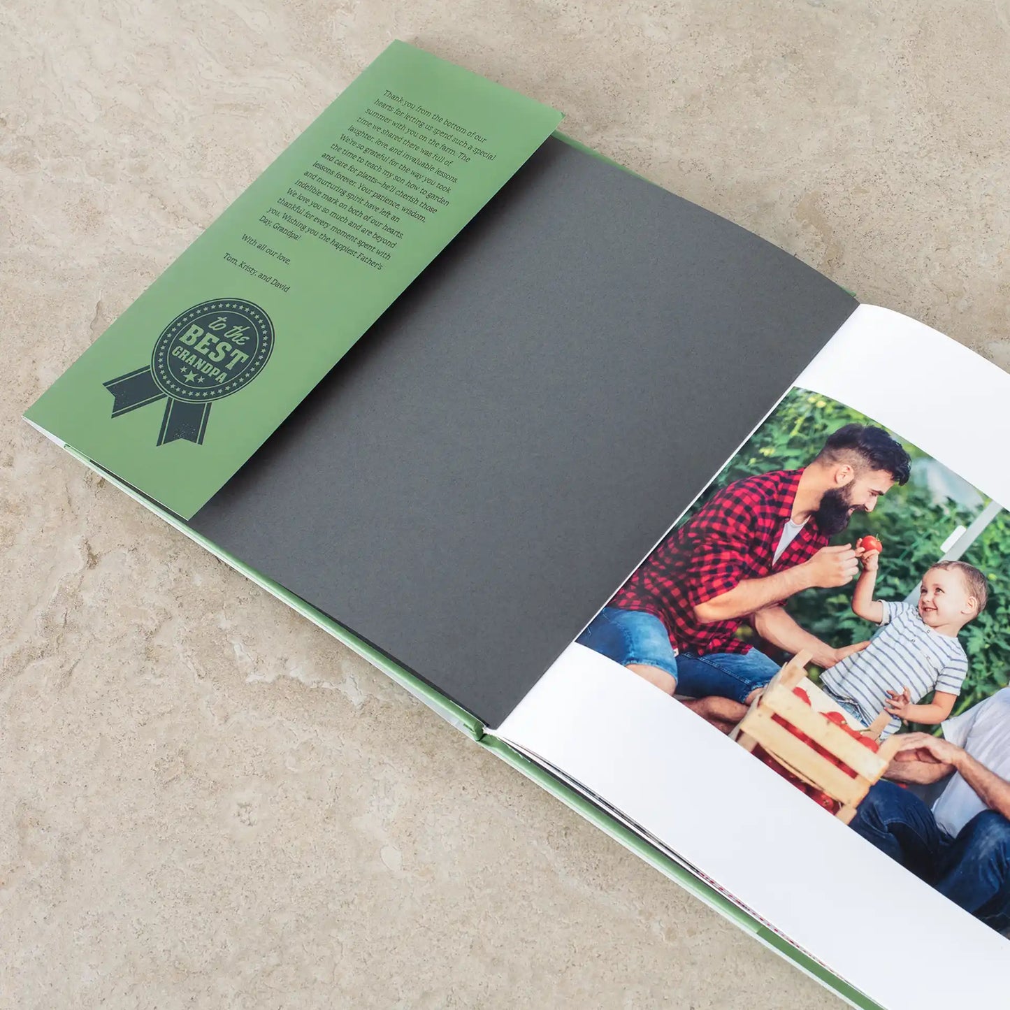 Open hardcover photo book showing a green interior page with a 'Best Grandpa' badge and a heartfelt handwritten message, alongside a photo of a father and young son picking tomatoes together in a garden