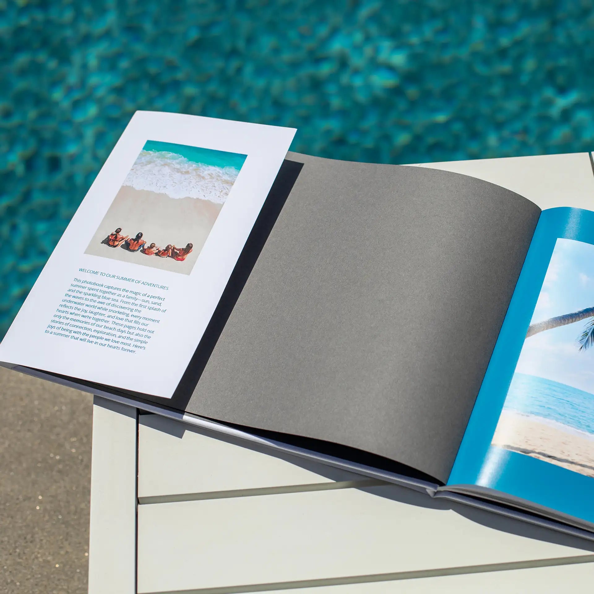 Hardcover photo book open to introductory page with small beach photo and printed text on left, opposite blank dark divider page near poolside.