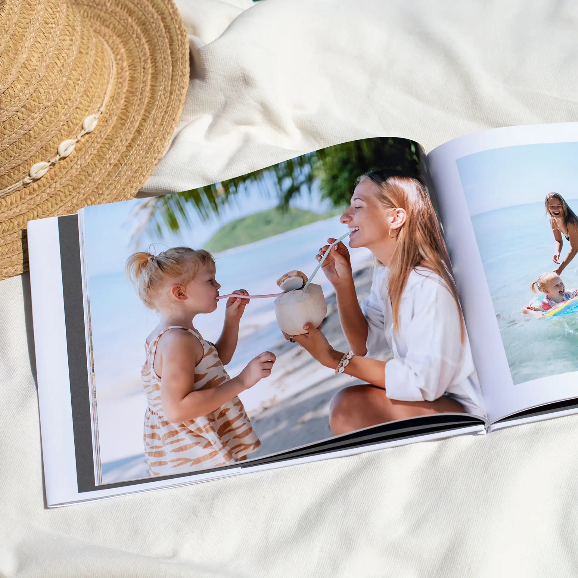 Hardcover photo book open to full-bleed beach image of adult and child drinking from coconut, resting on light fabric with sun hat.