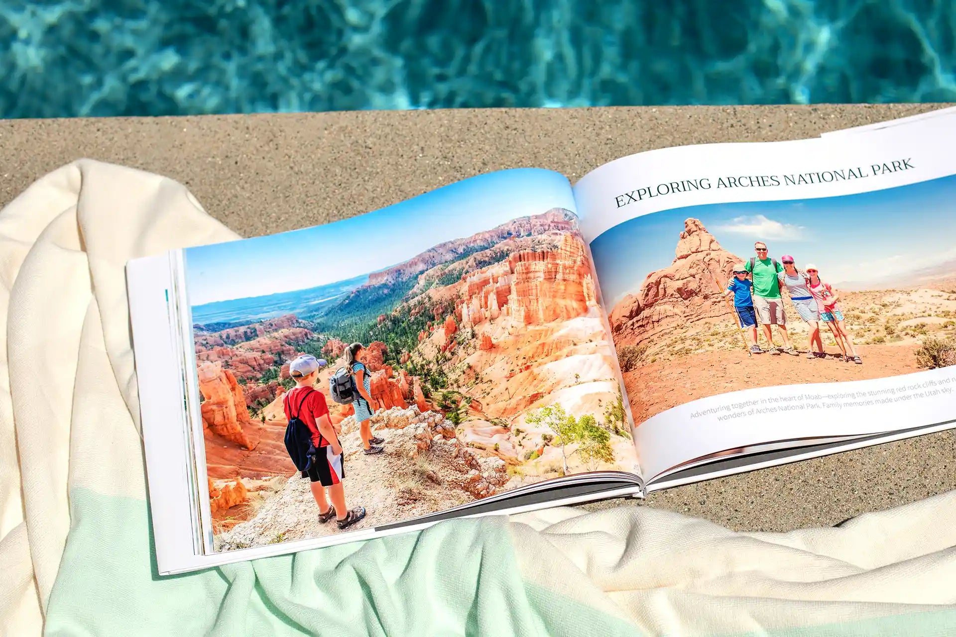 Open hardcover photo book resting on a towel beside a swimming pool, displaying a family travel spread titled 'Exploring Arches National Park' with a full-page landscape photo of children hiking among red rock formations and a family portrait posing together at the park