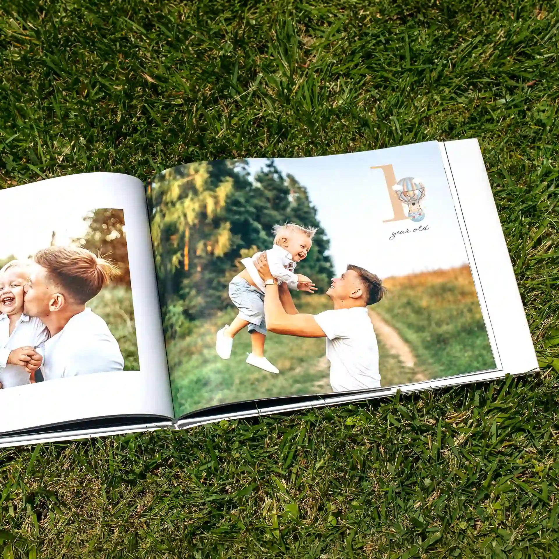 Open hardcover photo book lying on grass, displaying a "1 Year Old" milestone spread with a father lifting and laughing with his baby outdoors, alongside a smaller photo of the two sharing a joyful moment.