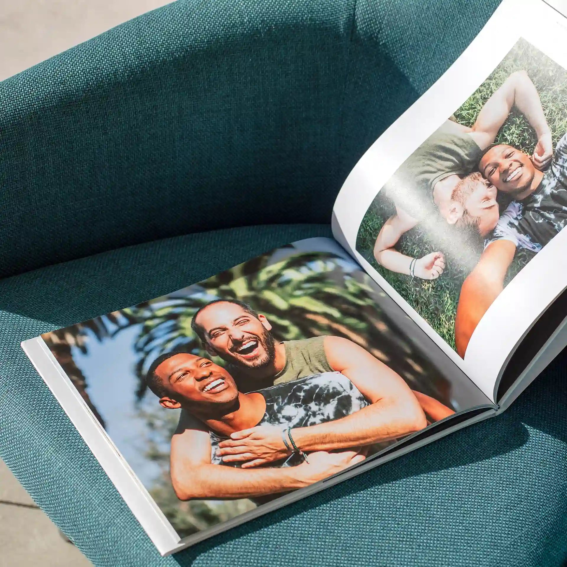 Open hardcover photo book resting on a teal armchair, showing photos of a couple embracing and laughing outdoors among palm trees, with an overhead shot of the couple lying together in grass on the facing page.