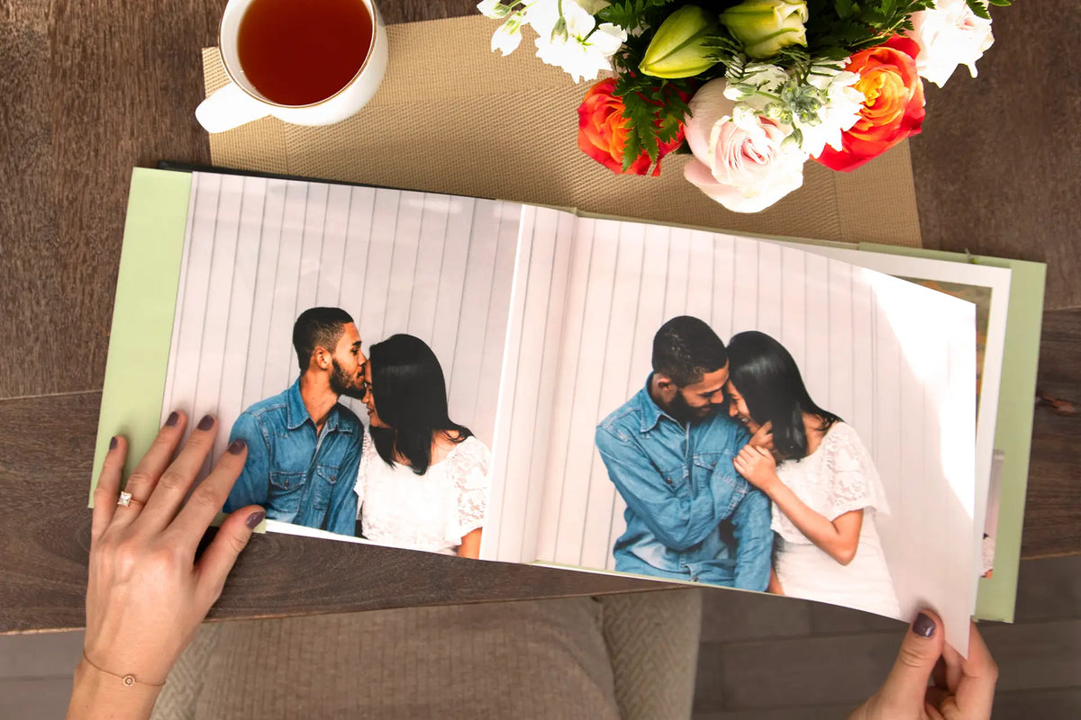 Hands turning the pages of an open hardcover photo book showing a smiling couple, beside a cup of tea and flowers.