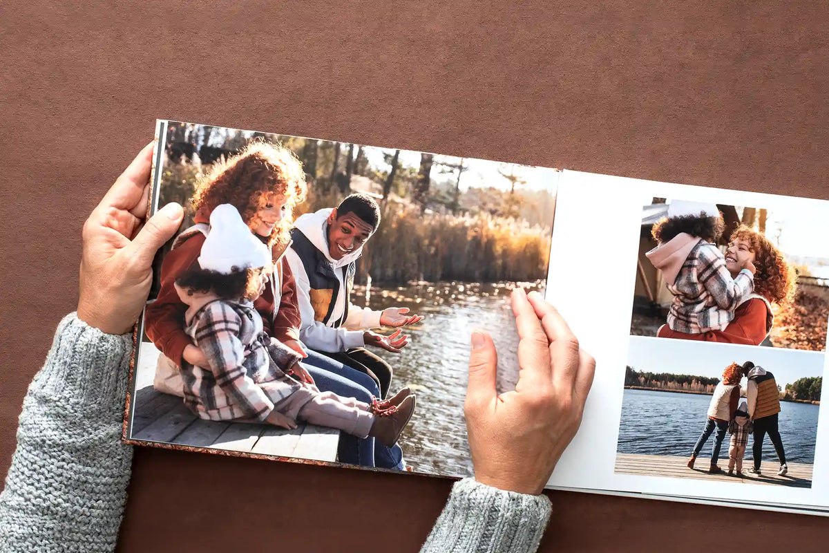 Hands turning the pages of a hardcover photo book open to a spread of a family on a lakeside dock in autumn, with a full page photo beside a two photo collage.
