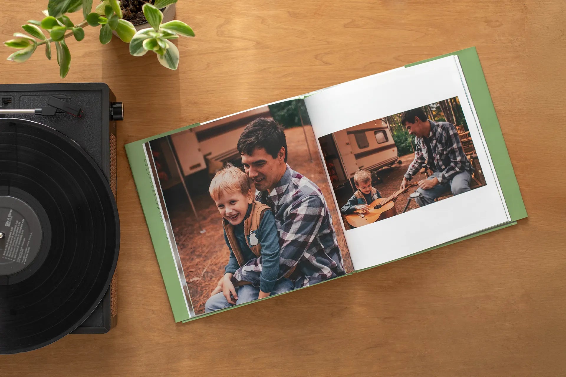 Open hardcover photo book resting on a wooden table beside a record player and houseplant, displaying a camping-themed spread with a large photo of a father and young son laughing together at a campsite, alongside a smaller photo of the boy learning to play guitar outdoors