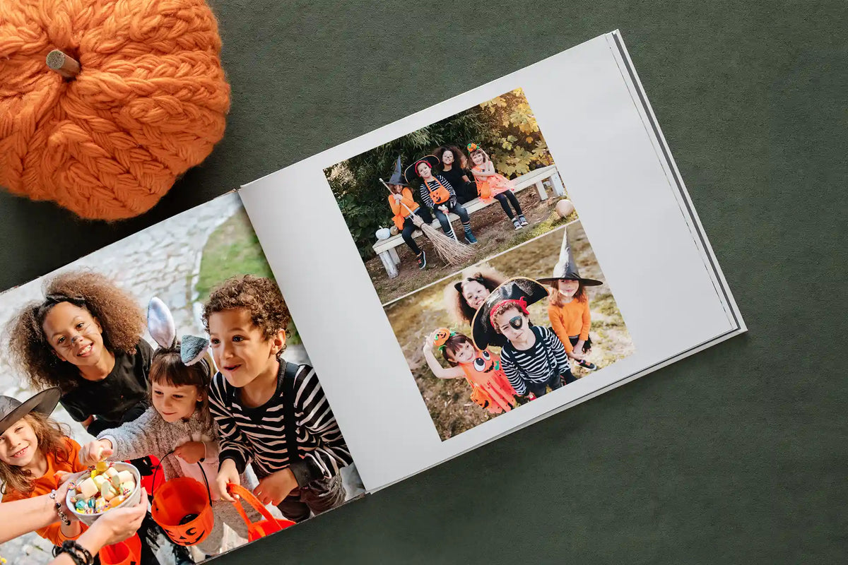 An open hardcover photo book with photos of costumed kids trick-or-treating and posing together, beside a knit orange pumpkin.
