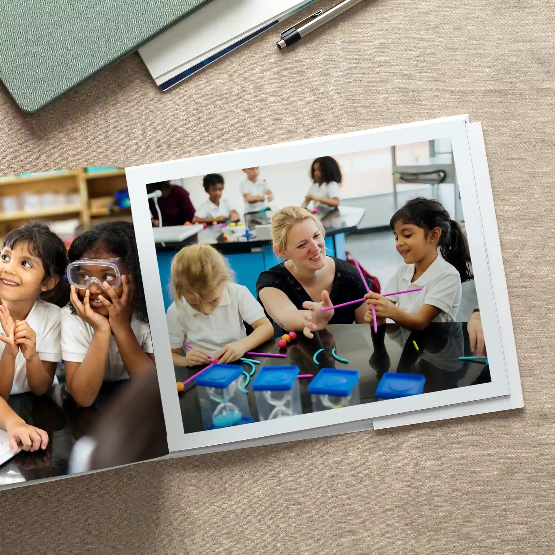 Open hardcover photo book showing school-themed pages: children in safety goggles during a science experiment on the left, and a teacher engaging students with colorful building materials on the right