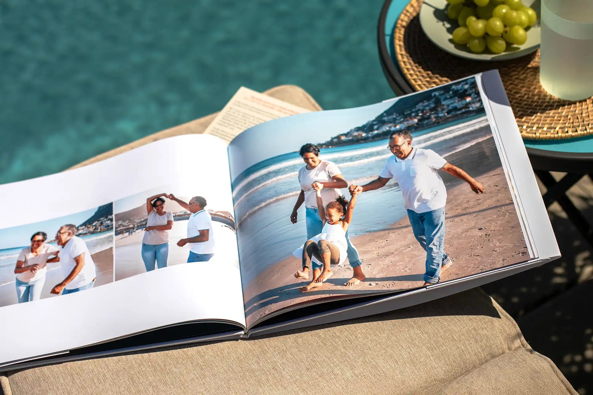 Open hardcover photo book on a lounge chair by a pool, displaying a large beach photo of two grandparents swinging a laughing child between them, with smaller photos of the couple dancing on the beach on the facing page.