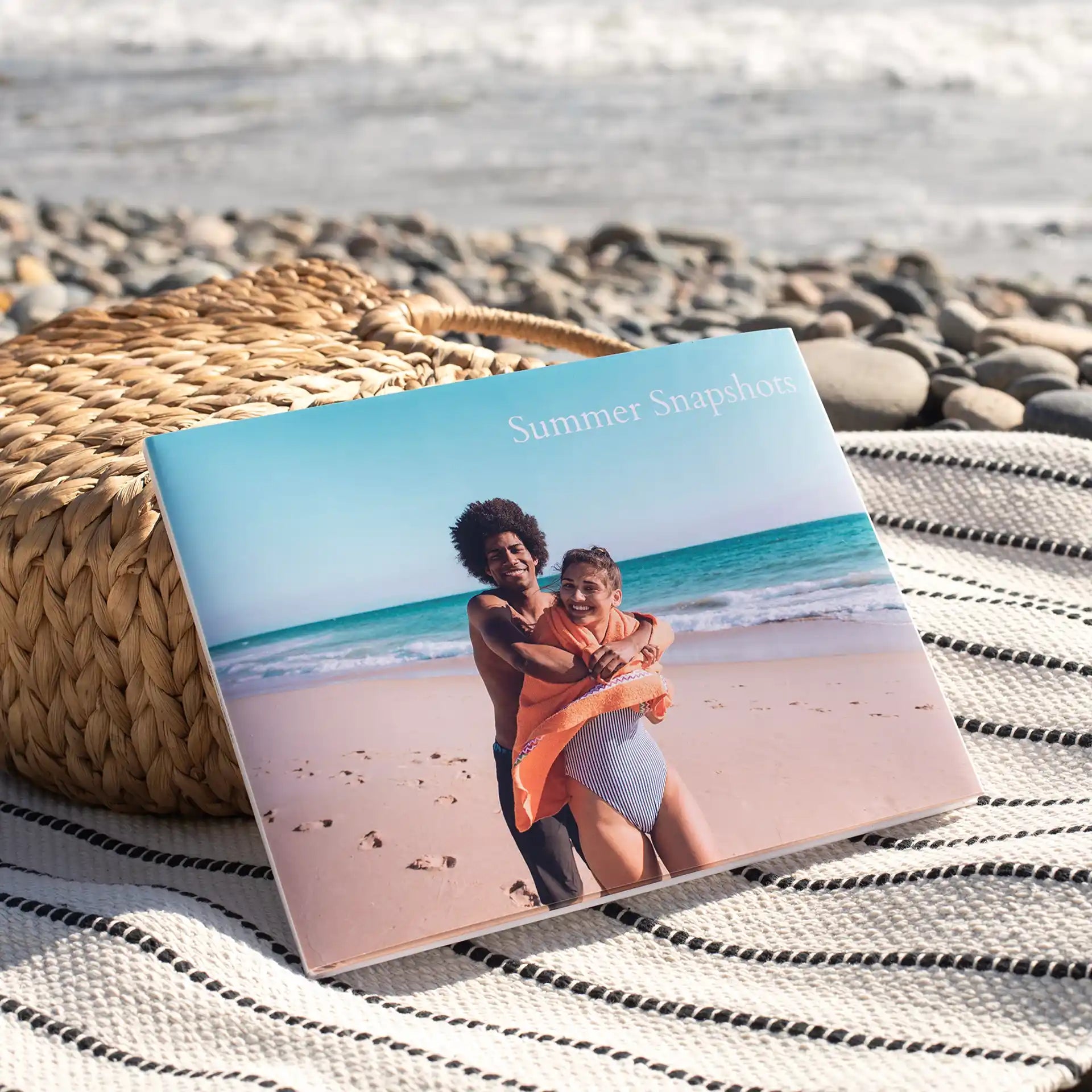 Closed hardcover photo book with beach couple image and “Summer Snapshots” title on cover, resting on woven blanket near rocky shoreline.