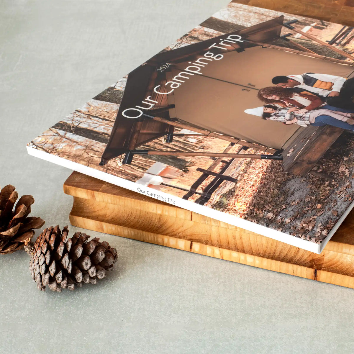 Closed hardcover photo book titled 'Our Camping Trip' with a full-cover family photo, resting on a wood board with pinecones.
