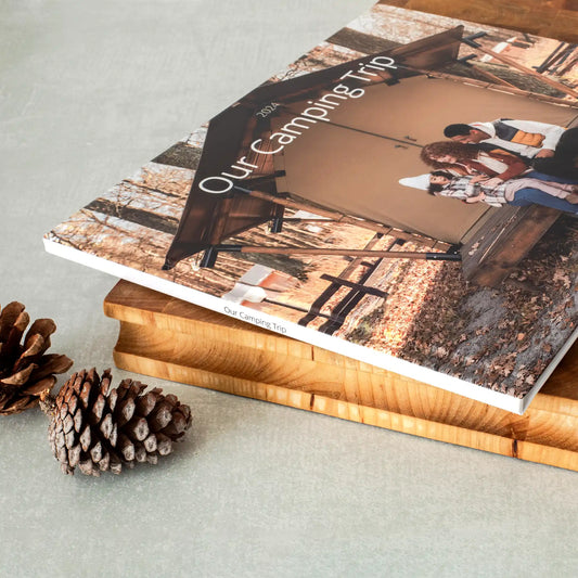 Closed hardcover photo book titled 'Our Camping Trip' with a full-cover family photo, resting on a wood board with pinecones.