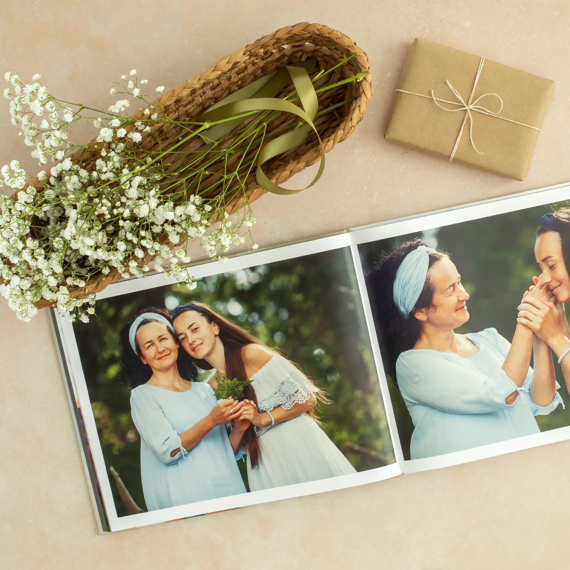 Hardcover photo book open to two-page spread with portraits of two people outdoors, styled with flowers, ribbon, and wrapped gift on a soft surface.