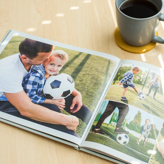 Open hardcover photo book displaying a full-page father and child soccer portrait with additional action photos on the right page, resting on a wooden table beside a coffee mug.