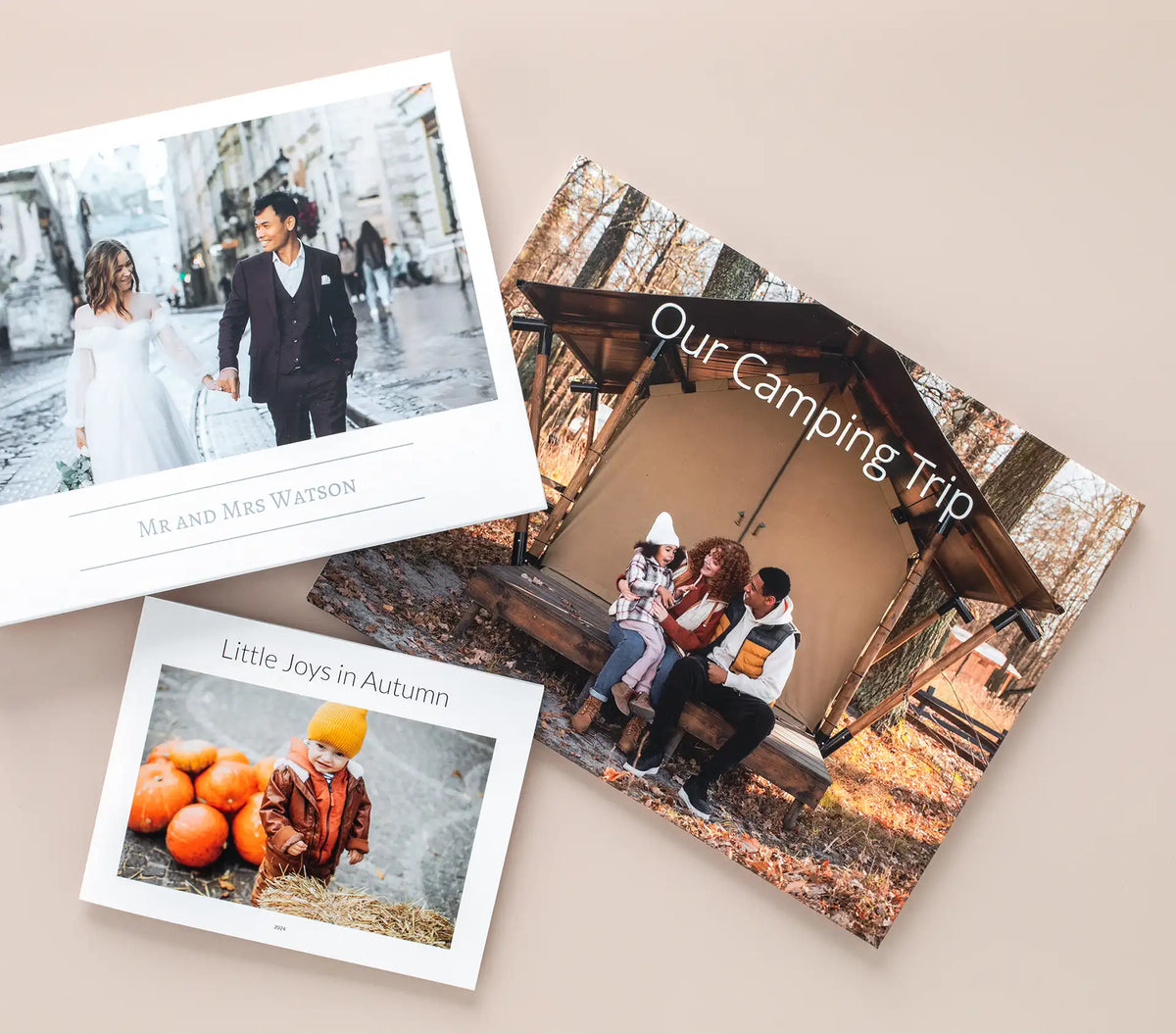 Three custom photo book covers fanned out — a wedding book titled 'Mr and Mrs Watson,' a family camping book titled 'Our Camping Trip,' and a children's book titled 'Little Joys in Autumn.