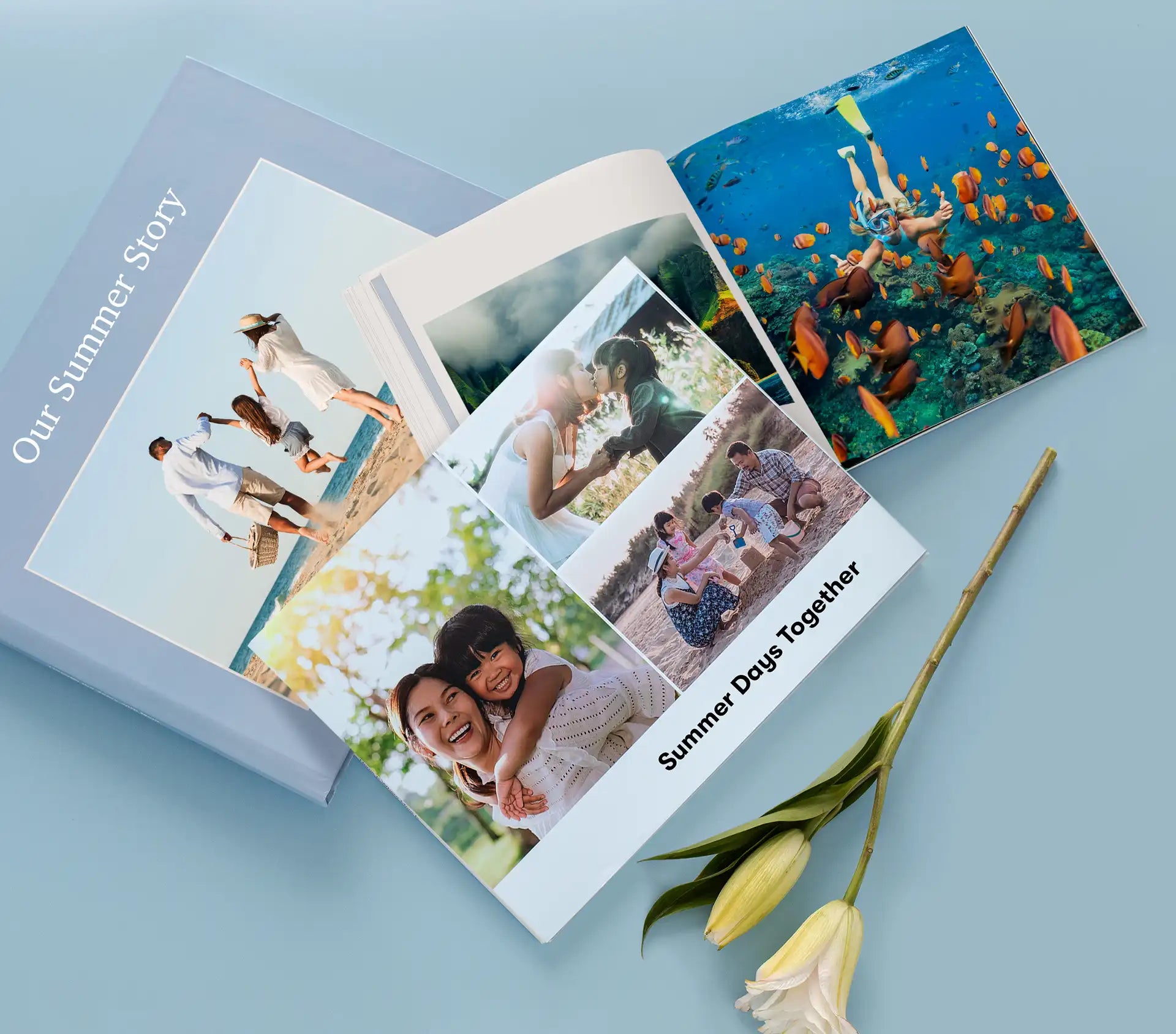 Assorted photo books displayed on a soft blue surface, featuring vibrant covers and open pages with family moments, travel scenes, and underwater photography, alongside a white lily flower.