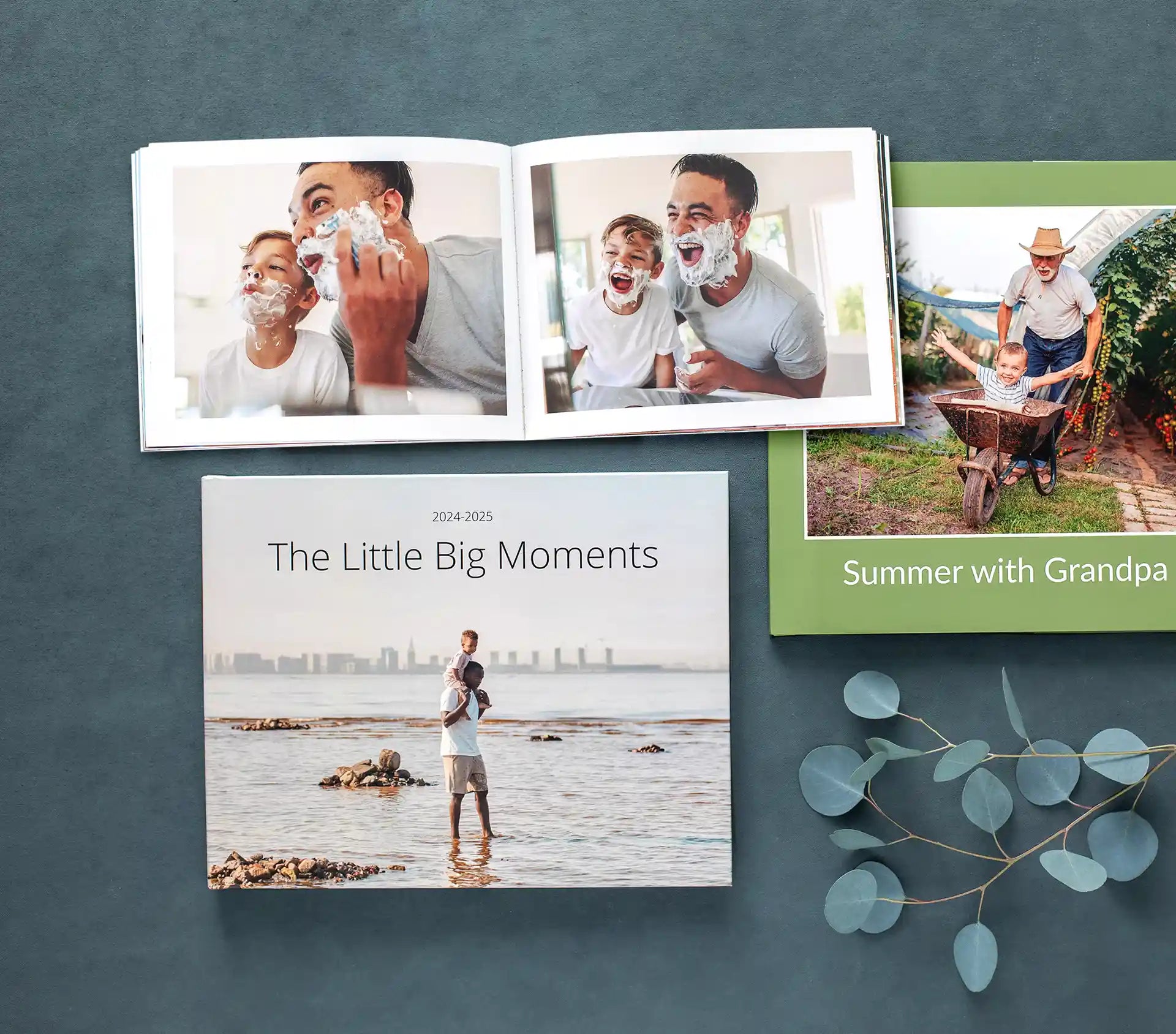 A collection of three custom photo books displayed on a dark surface with eucalyptus stems, featuring an open layflat book showing a father and son shaving together, a hardcover titled 'Summer with Grandpa,' and a softcover titled 'The Little Big Moments