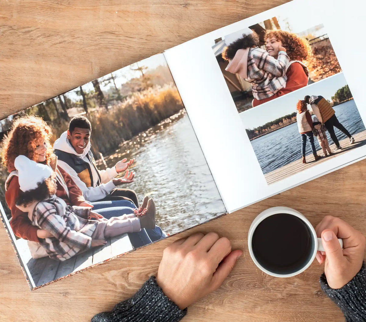 Hardcover photo book open on a wooden table to full-spread photos of a family relaxing by a lake in autumn, with a cup of coffee nearby.