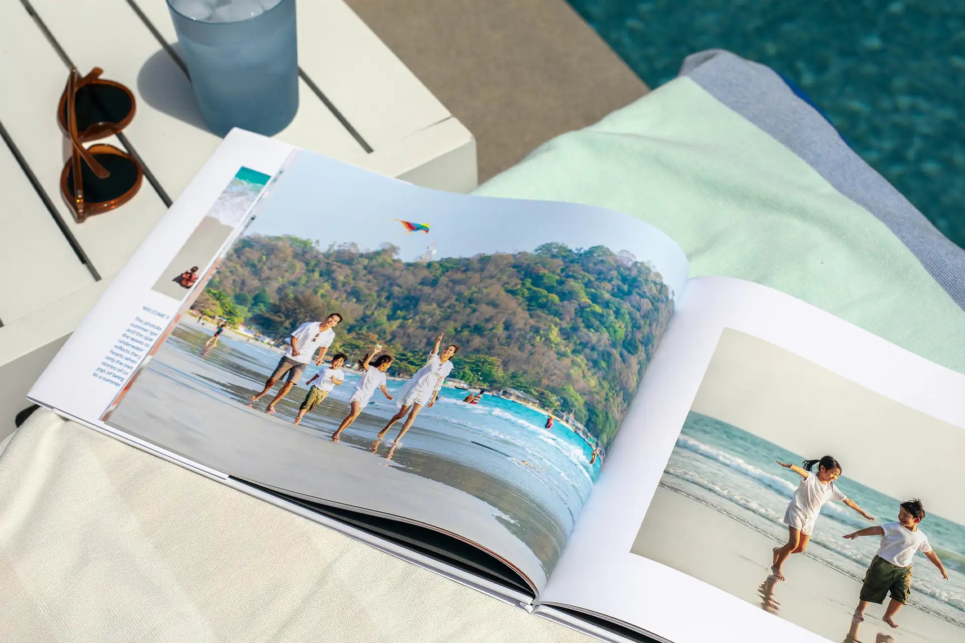 Hardcover photo book open to bright beach scenes of children and family playing by the ocean, placed near a pool.