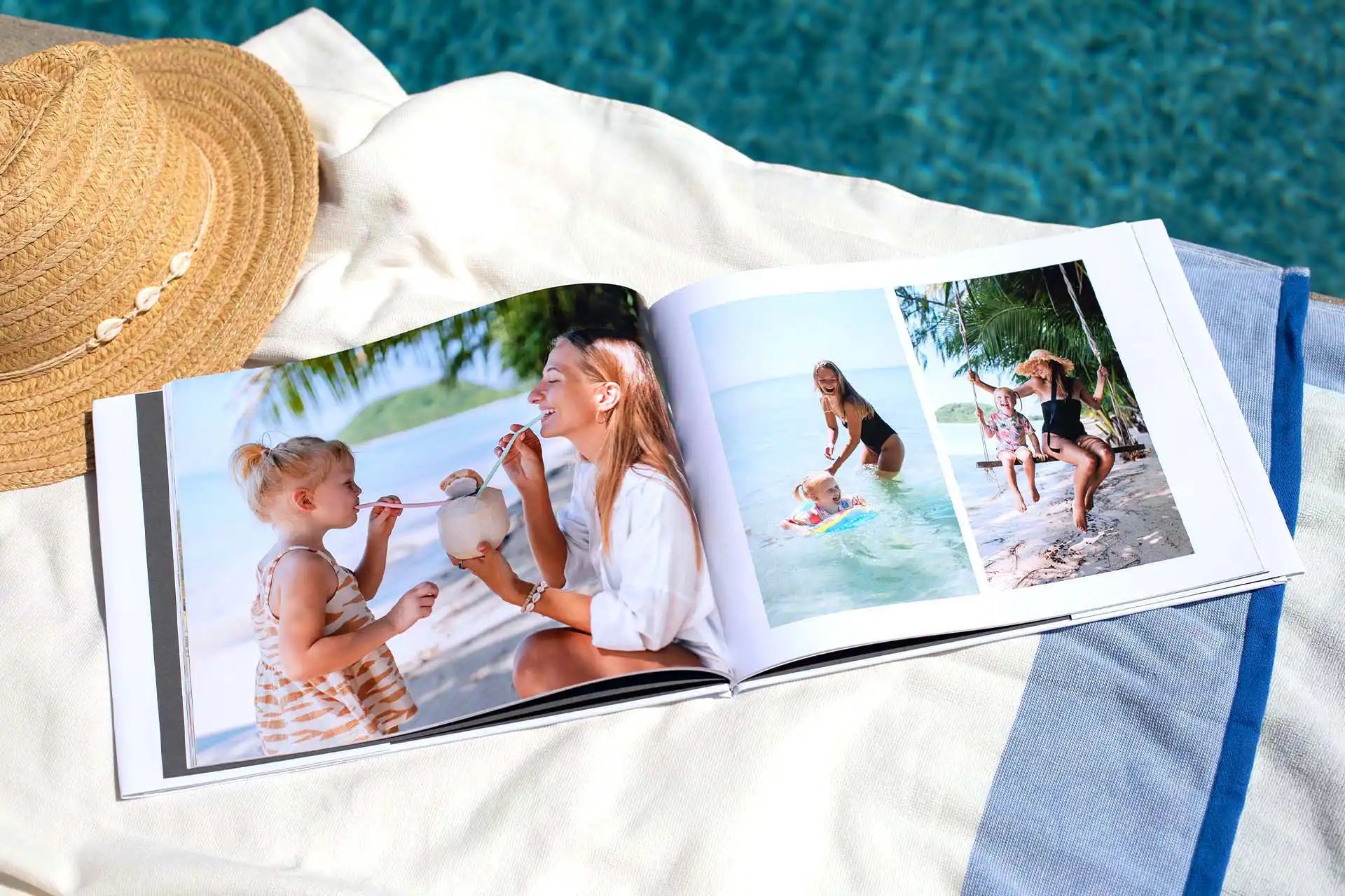 Open hardcover photo book displayed poolside on towel, showing full-bleed family beach photos across panoramic layflat pages.