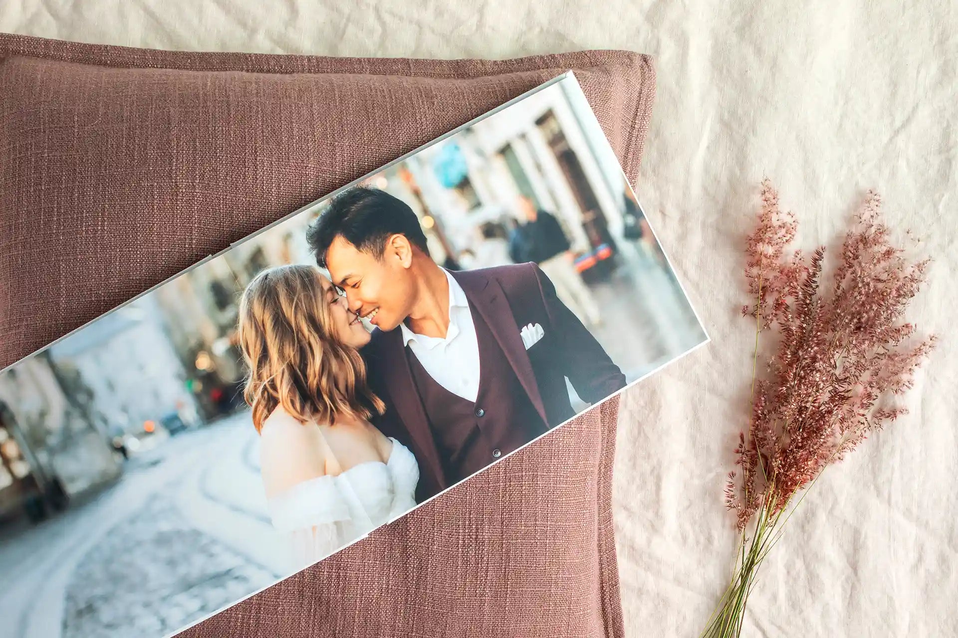Open layflat photo book resting on a mauve linen pillow, showing a full-spread photo of a bride and groom embracing on a city street, with dried pink flowers beside it.