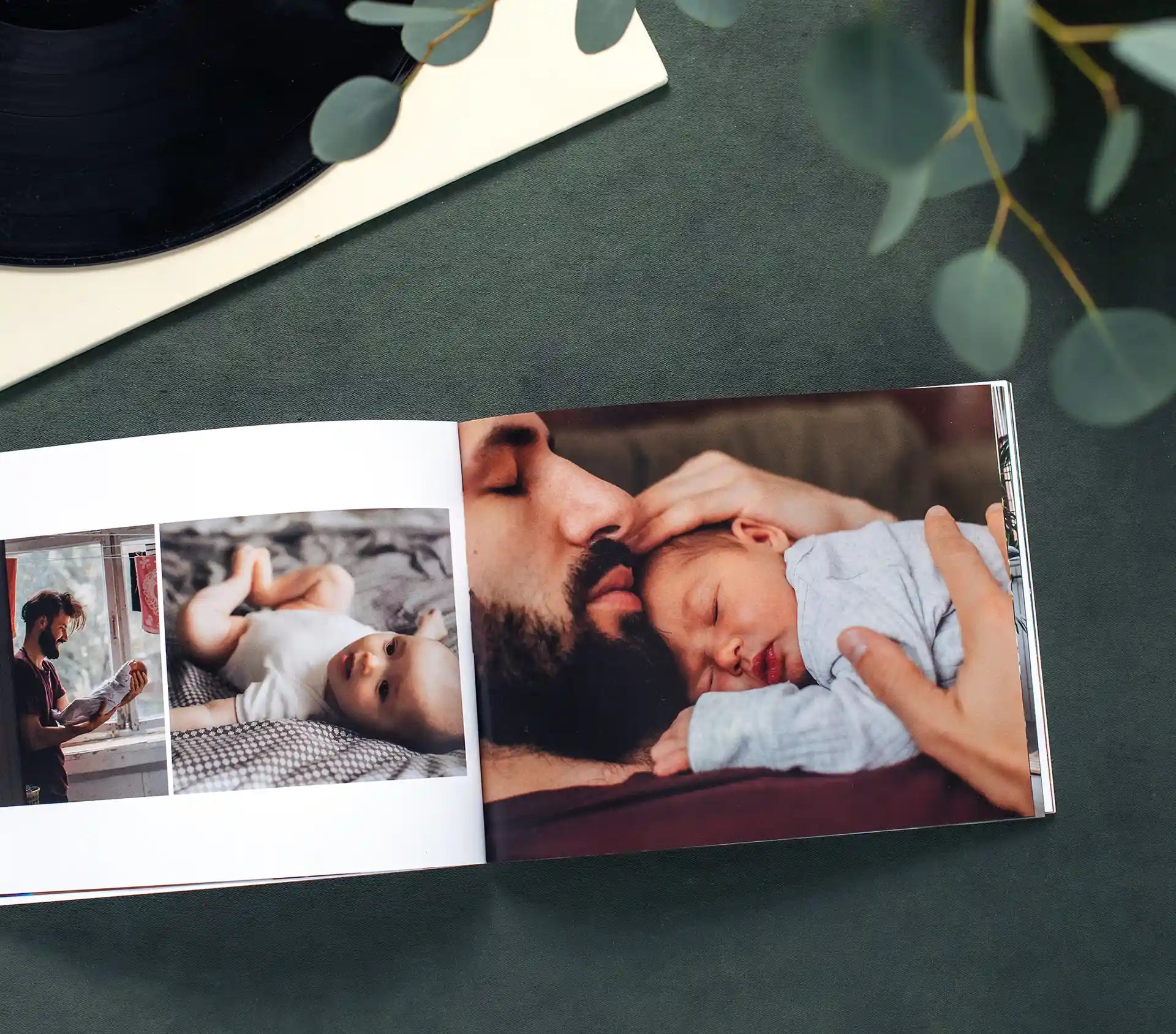Open softcover photo book on a dark green surface beside a vinyl record and eucalyptus stems, displaying a tender newborn spread with a collage of a father caring for his baby alongside a full-page close-up of a bearded father gently kissing his sleeping newborn