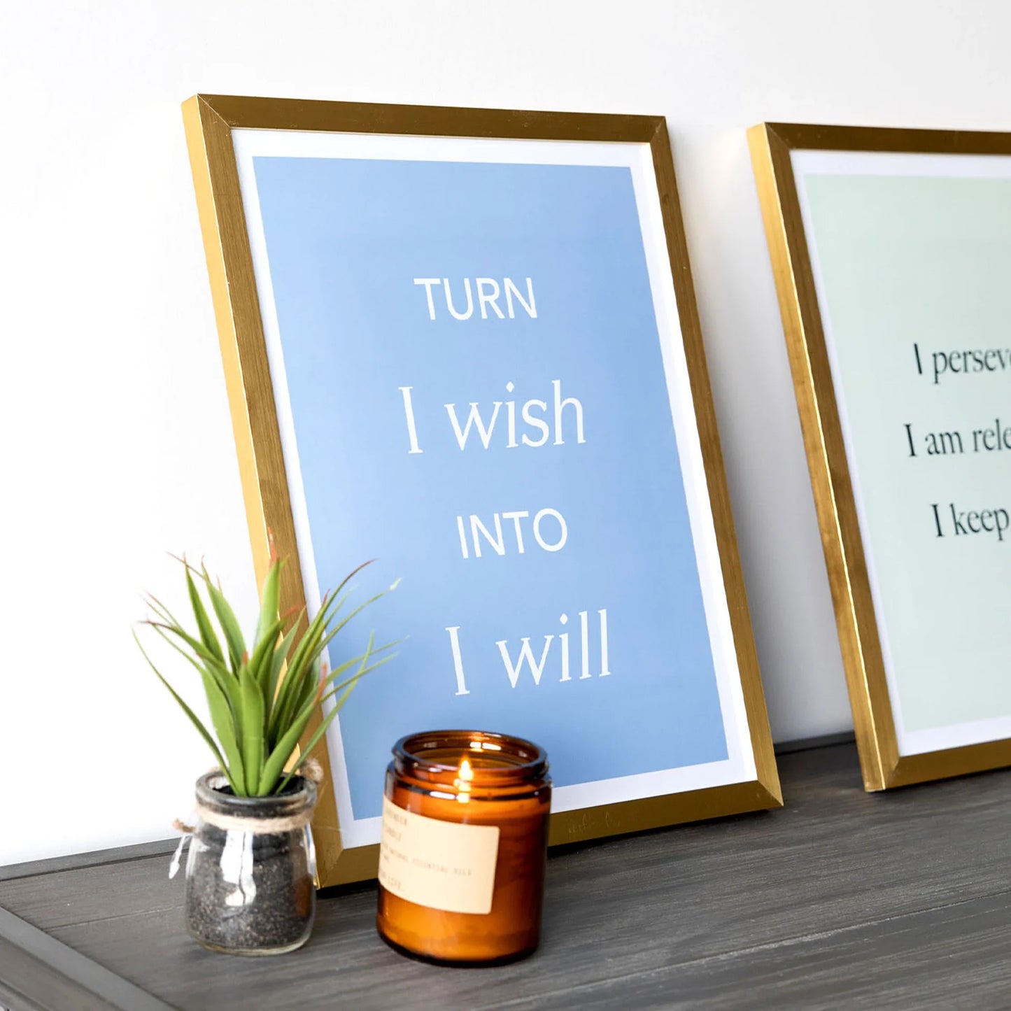 Gold framed large-format vertical typography print reading “Turn I wish into I will,” displayed on a dark tabletop beside a small plant and lit candle.