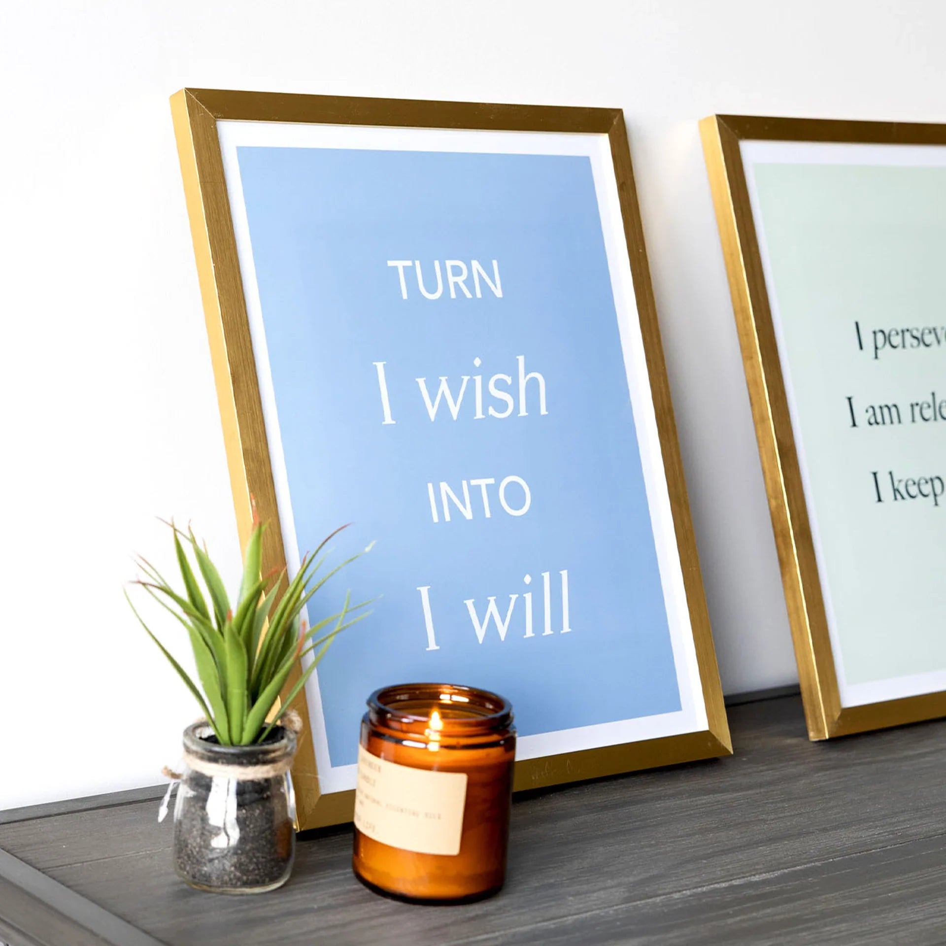 Gold framed large-format vertical typography print reading “Turn I wish into I will,” displayed on a dark tabletop beside a small plant and lit candle.